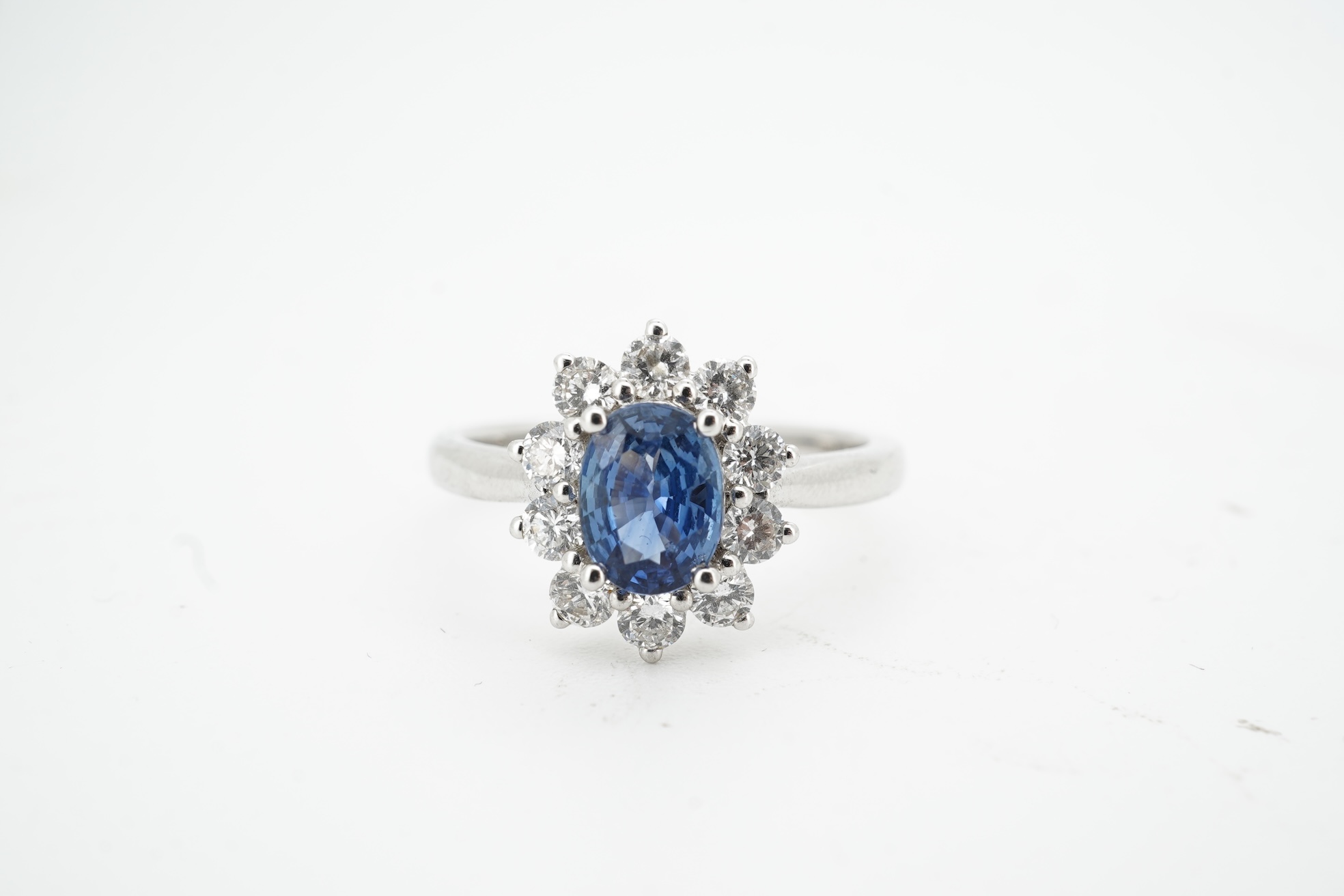 A sapphire and diamond cluster ring, claw-set with an oval sapphire weighing approximately 0.77 carats, within a border of brilliant-cut diamonds totalling approximately 0.50 carats, mounted in white gold, size M, stampe